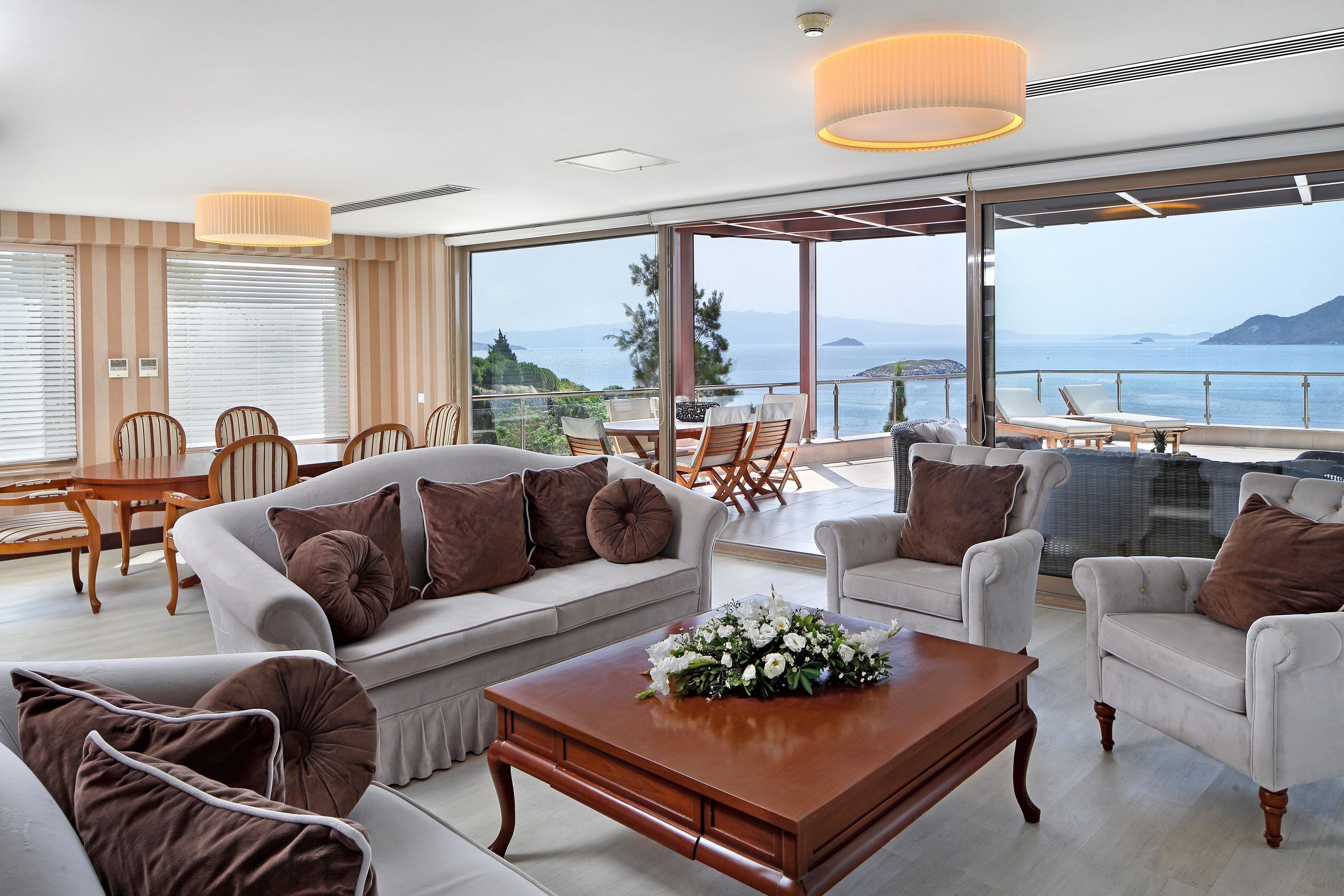 A modern living room with a large sofa, armchairs, a coffee table, and a dining area. It features large windows with a view of the sea and mountains.