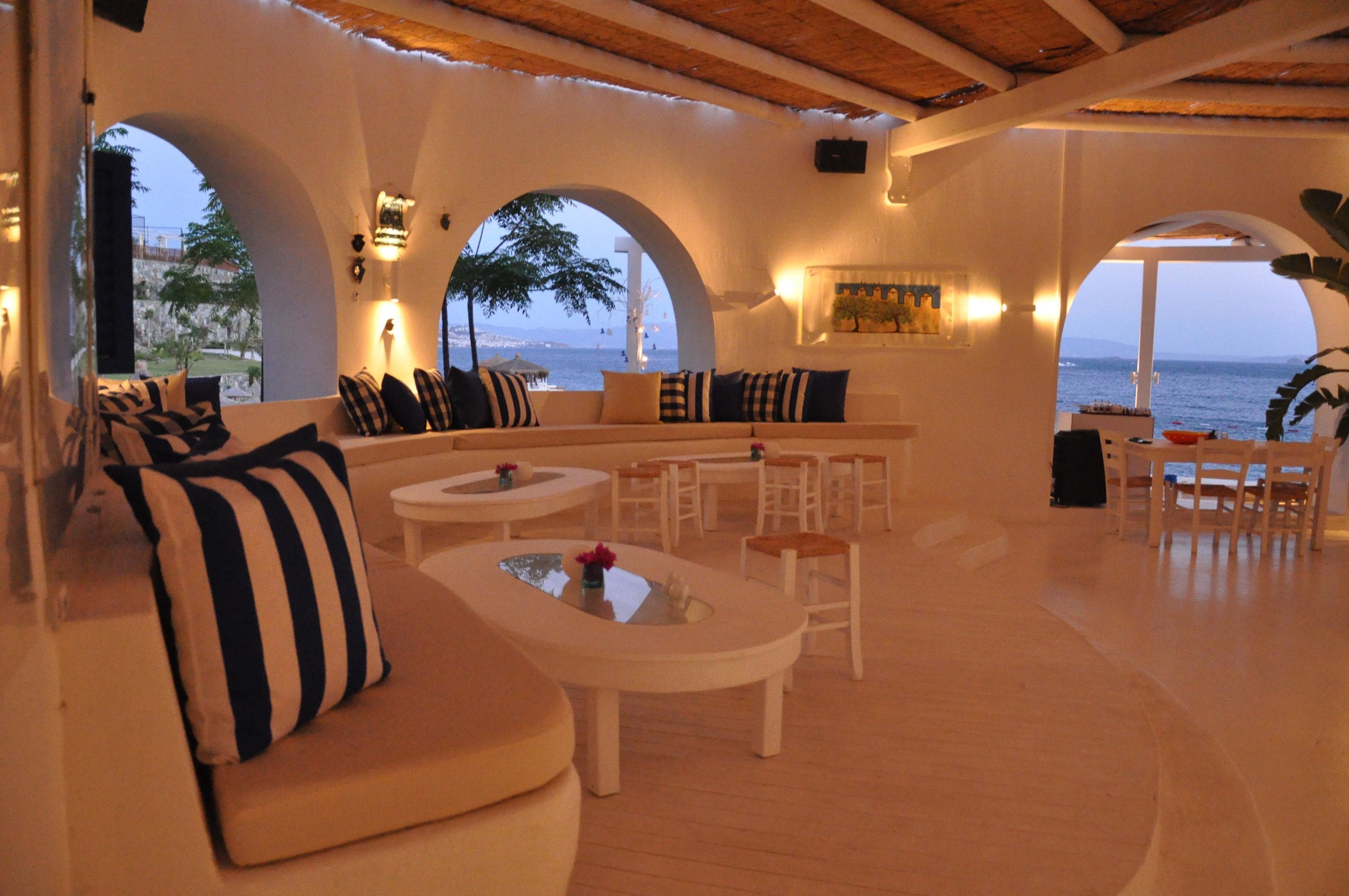 Un vaste espace de détente comprenant un canapé, des fauteuils et des tables, avec de grandes fenêtres en arc et une vue sur la mer.