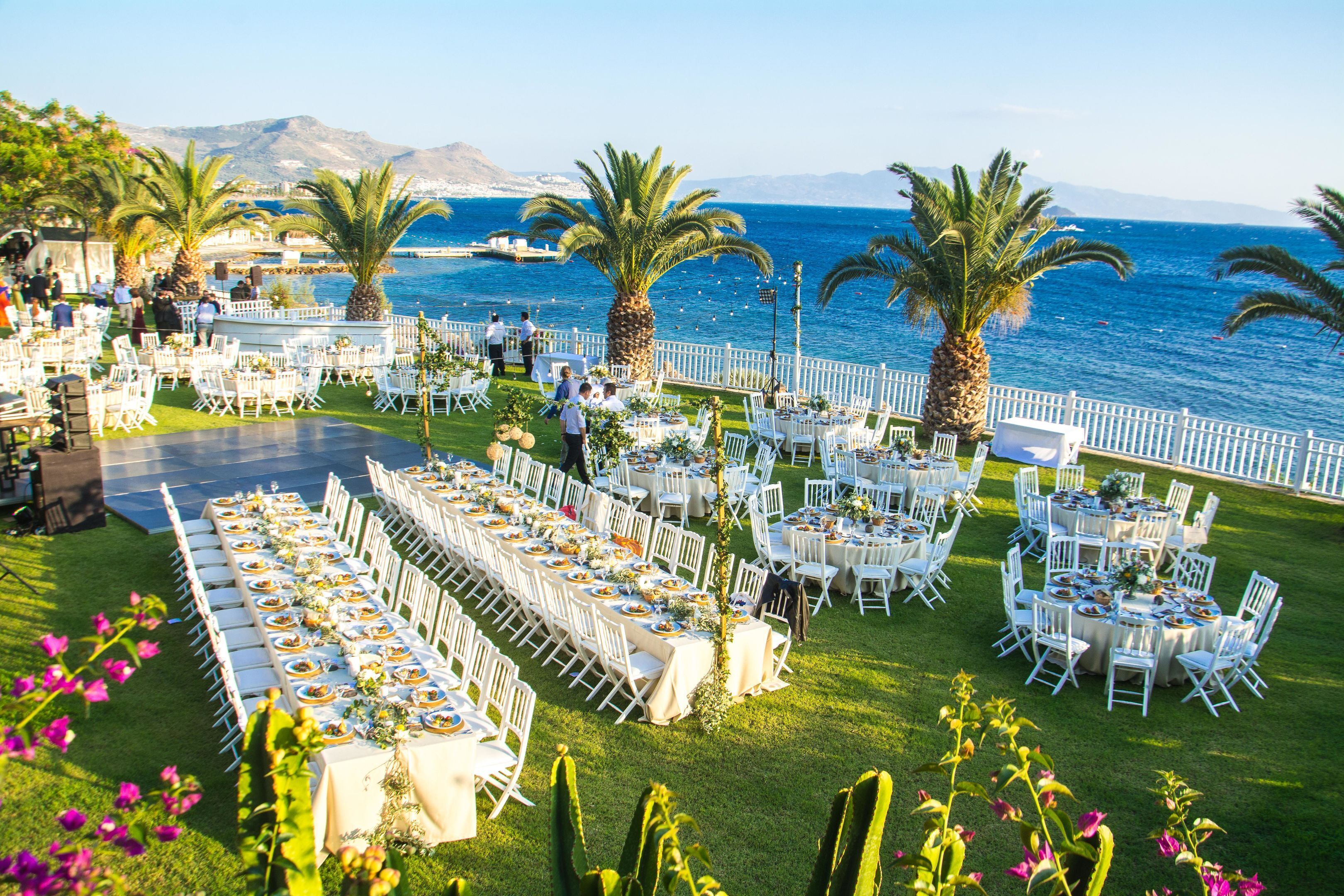 Een lange tafel gedekt voor een bruiloftsreceptie, met stoelen en tafels op een gazon met uitzicht op zee.