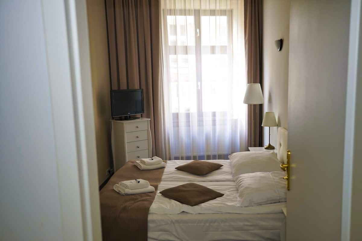 A hotel room with a bed, pillows, a TV, a lamp, and a window with curtains.