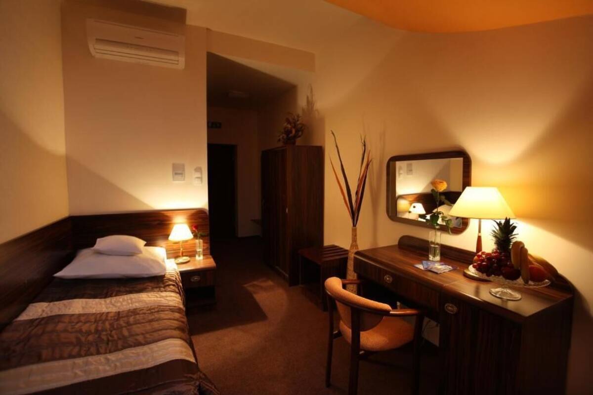 A hotel room with a bed, a desk with a lamp, a chair, a mirror, and a fruit bowl.