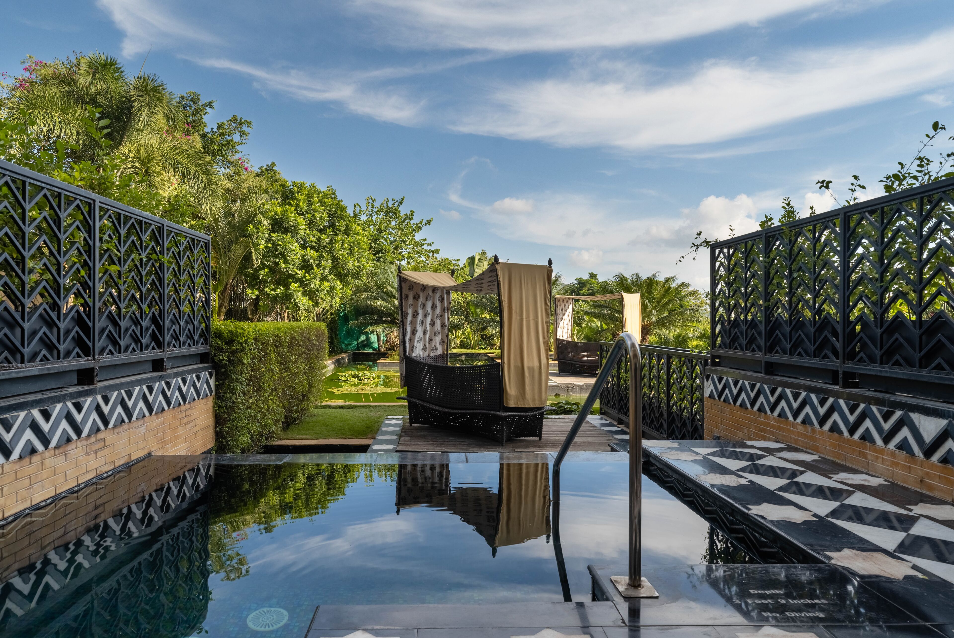 A pool area with a black and white checkered floor, a black wicker chair with a beige canopy, and a metal fence with a zigzag pattern.
