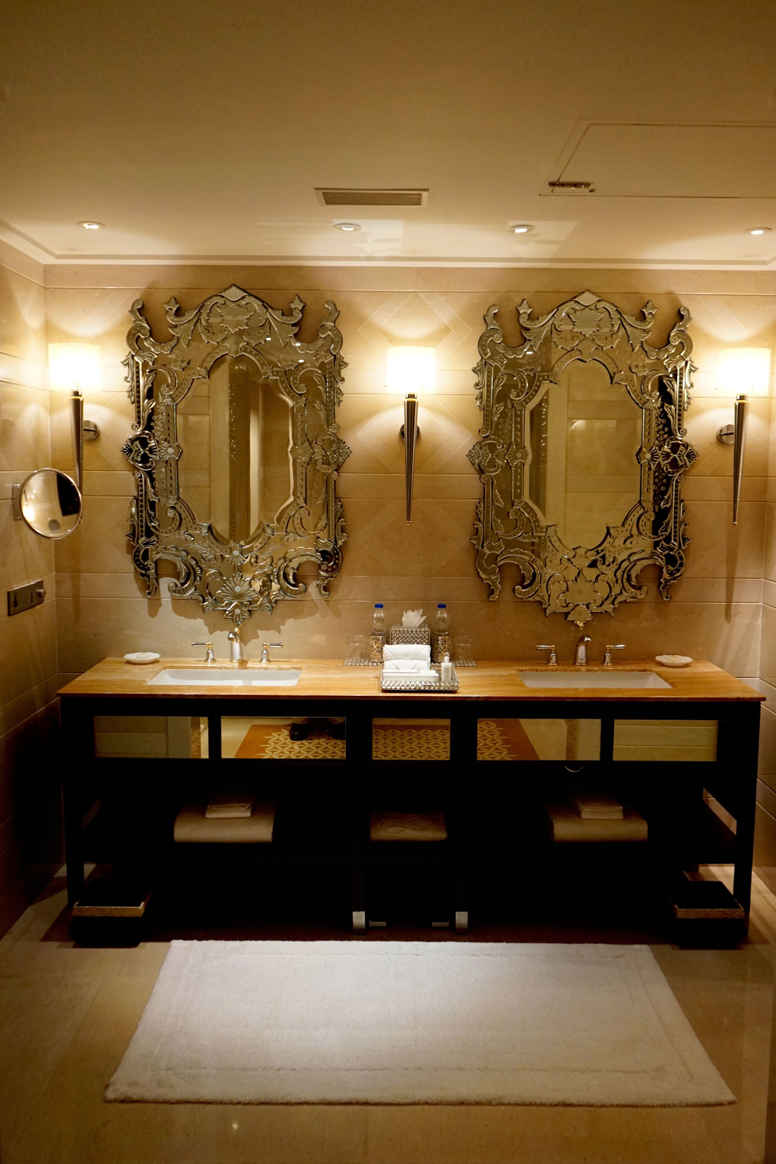 A bathroom with two large ornate mirrors, a wooden vanity, and a white rug.