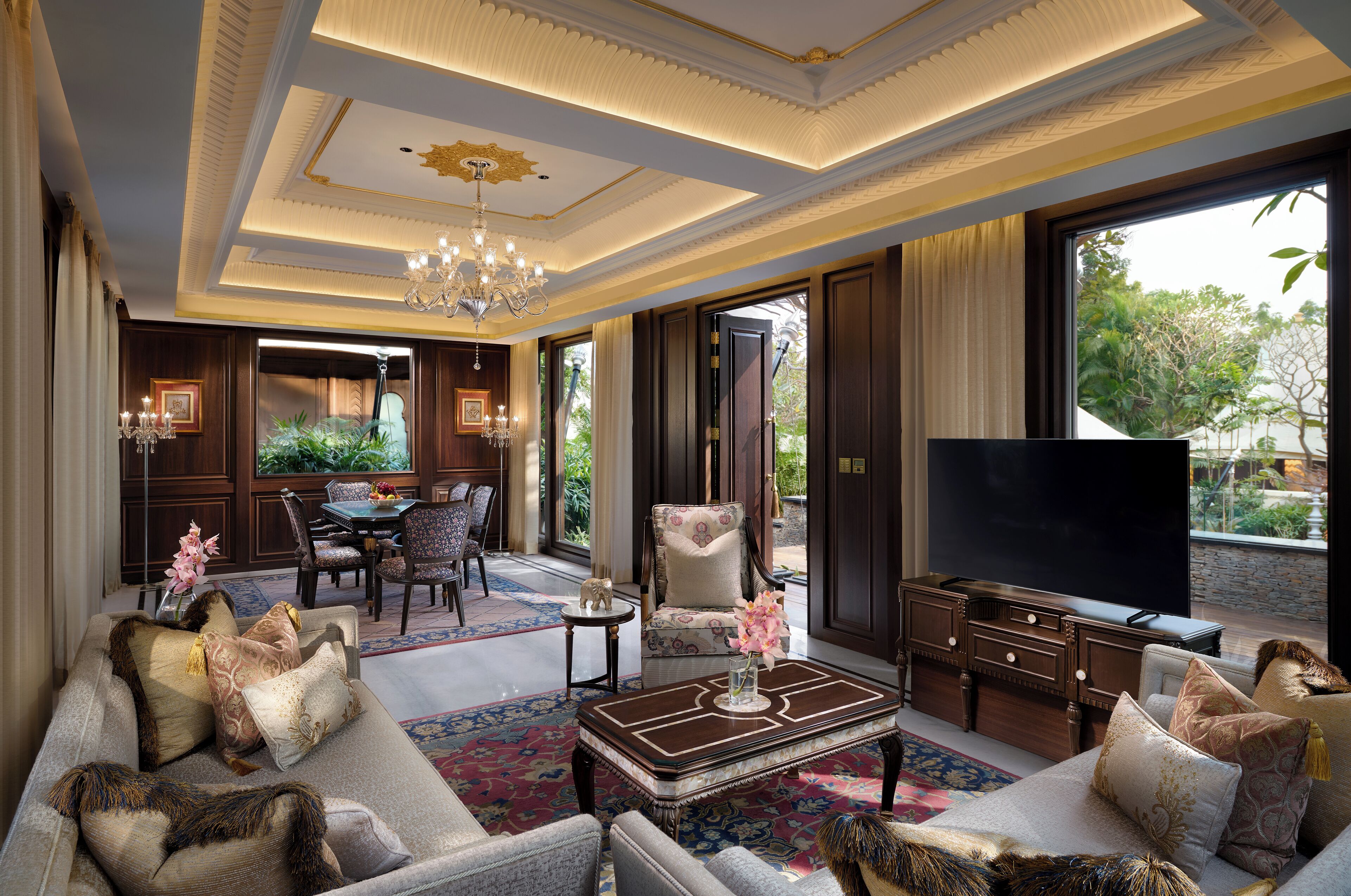 A spacious living room with a large sofa, a coffee table, and a television. There is a dining area with a table and chairs, and a chandelier hanging from the ceiling.