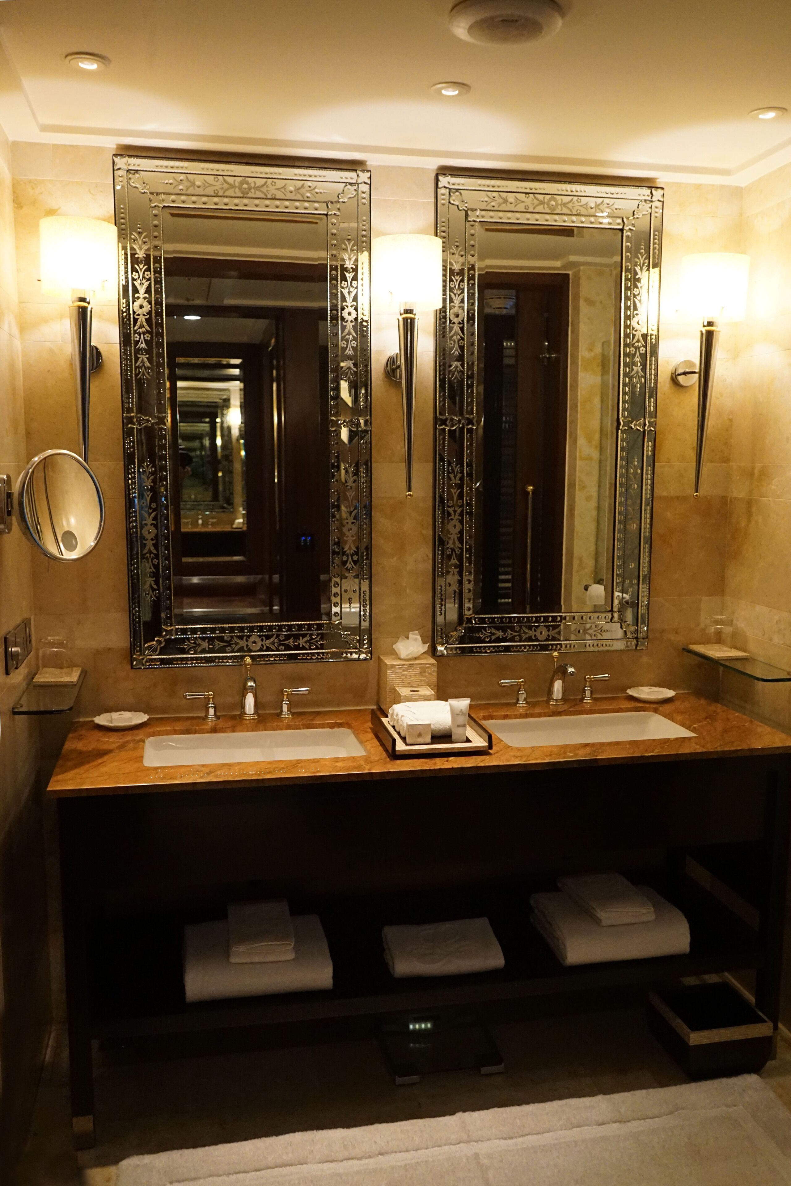 A bathroom with two sinks, two large mirrors, and wall sconces. There are towels and toiletries on the counter.