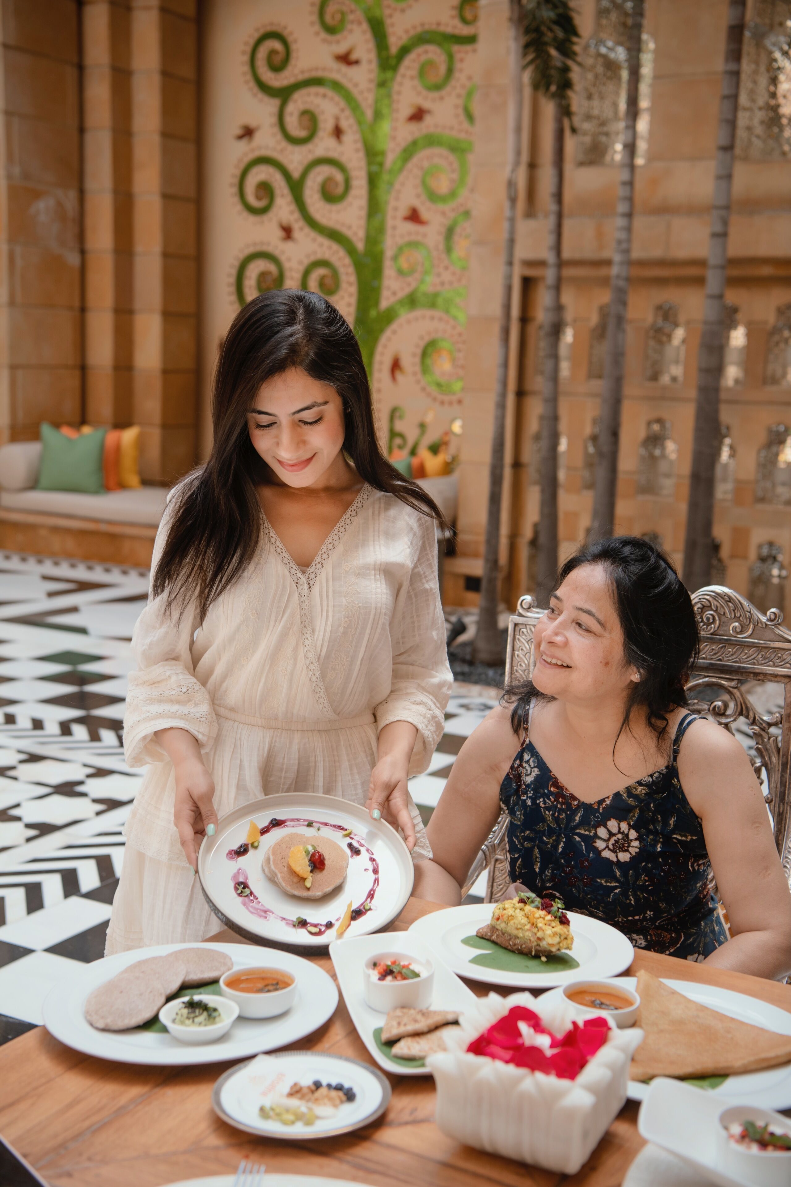 A dining area with a table set for two, featuring a variety of dishes and a decorative wall with a tree design.