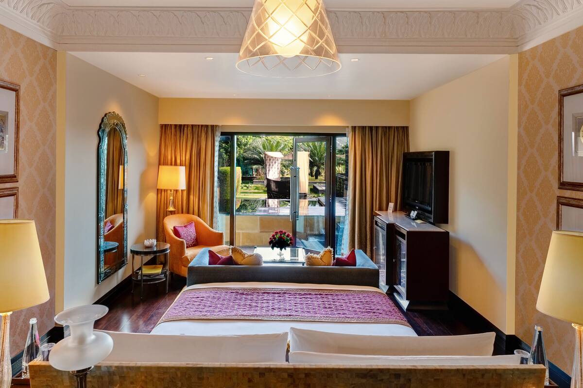 Ein Hotelzimmer mit einem großen Bett, einem Fernseher, einem Sofa, einem kleinen Tisch mit Blumen und Blick ins Freie.