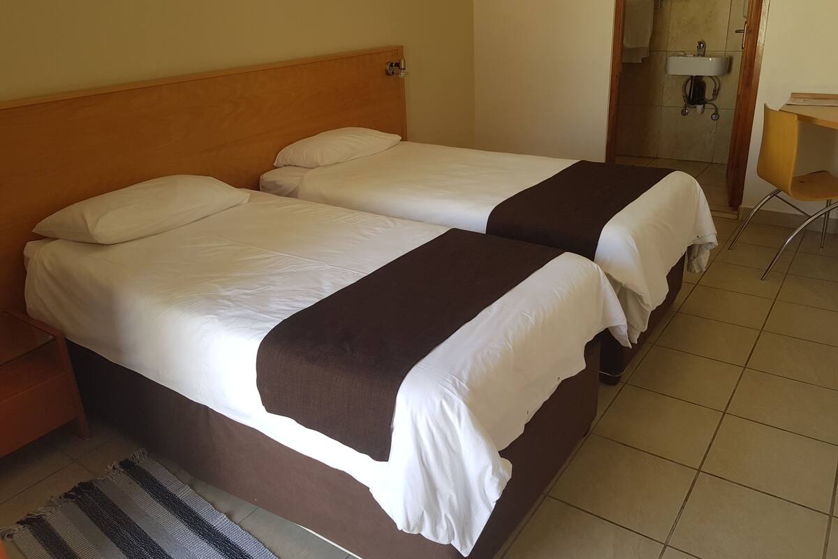 A room with two beds, a wooden headboard, a small desk, and a bathroom visible through an open door.