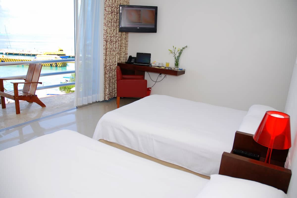 A hotel room with two beds, a desk with a computer, a TV, and a view of the sea.