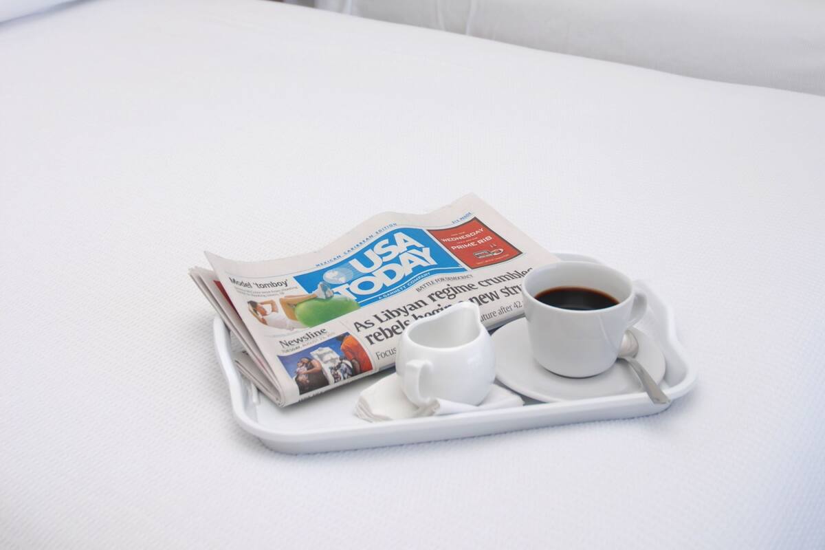 A hotel room with a bed, a nightstand with a red lamp, and a tray with a newspaper, coffee cup, and saucer.