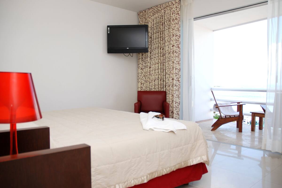 A hotel room with a bed, a TV mounted on the wall, a red chair, a small table, and a view of the ocean through a large window.