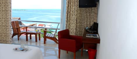 A hotel room with a balcony overlooking the sea, a bed, a desk with a chair, and a TV mounted on the wall.