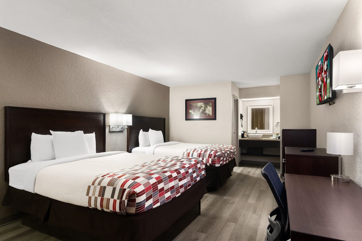 A hotel room with two beds, a desk, a chair, a TV, and a bathroom visible through an open door.