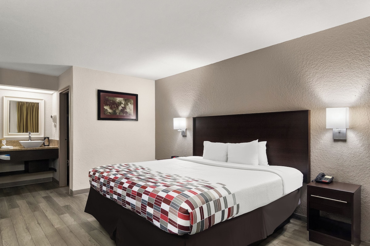 A hotel room with a large bed, a bedside table with a phone, a nightstand, a wall-mounted lamp, a framed picture, and a bathroom with a sink and mirror.