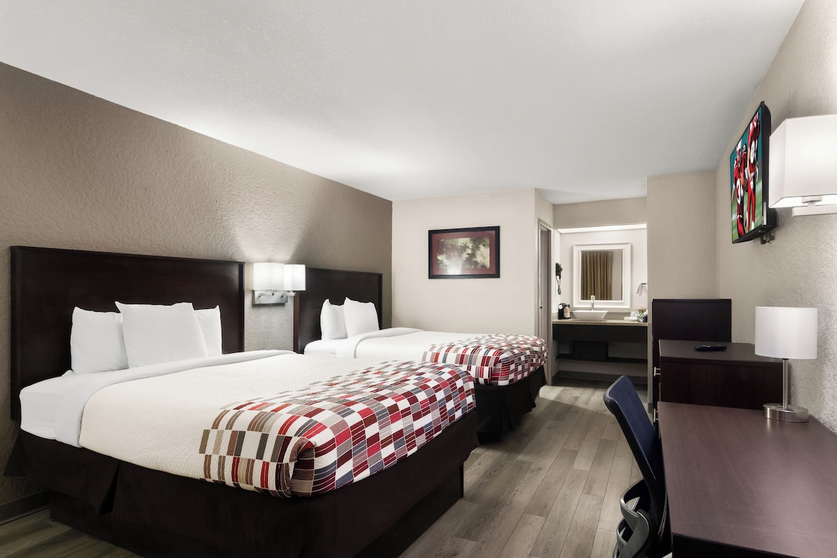 A hotel room with two beds, a desk, a chair, a TV, and a bathroom visible through an open door.