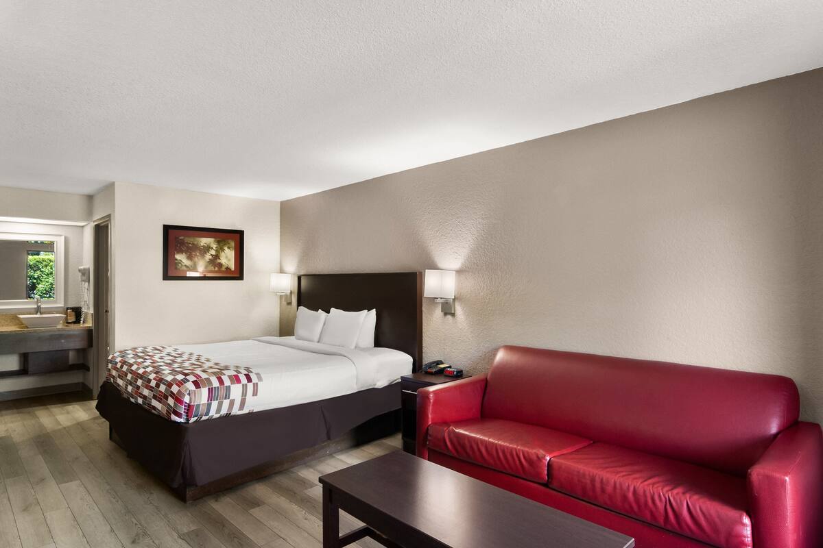A hotel room with a bed, a red leather sofa, a small table, a nightstand, a wall-mounted lamp, and a painting on the wall.
