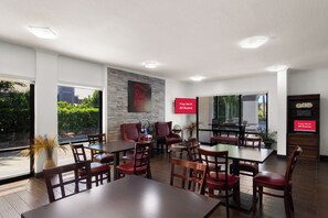 A hotel dining area with multiple tables and chairs, a wall-mounted TV displaying 'Free Wi-Fi All Rooms', and a view of greenery outside. - Red Roof Inn Kingsland (Kingsland)