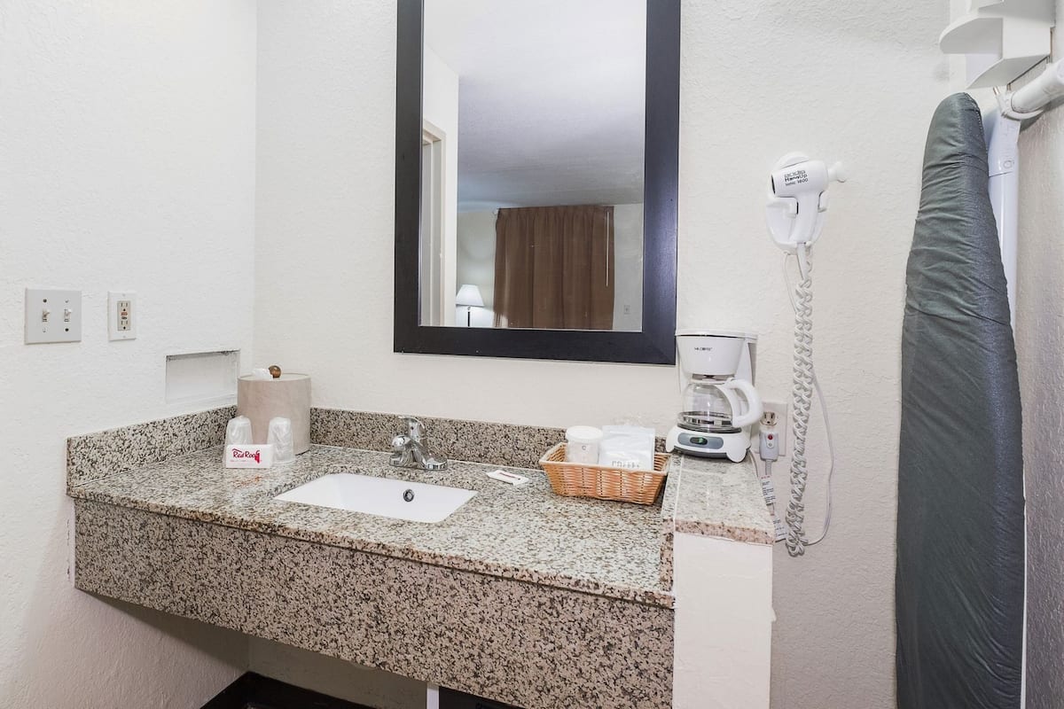 A bathroom with a granite countertop, a sink, a mirror, a hairdryer, a coffee maker, and a towel rack.