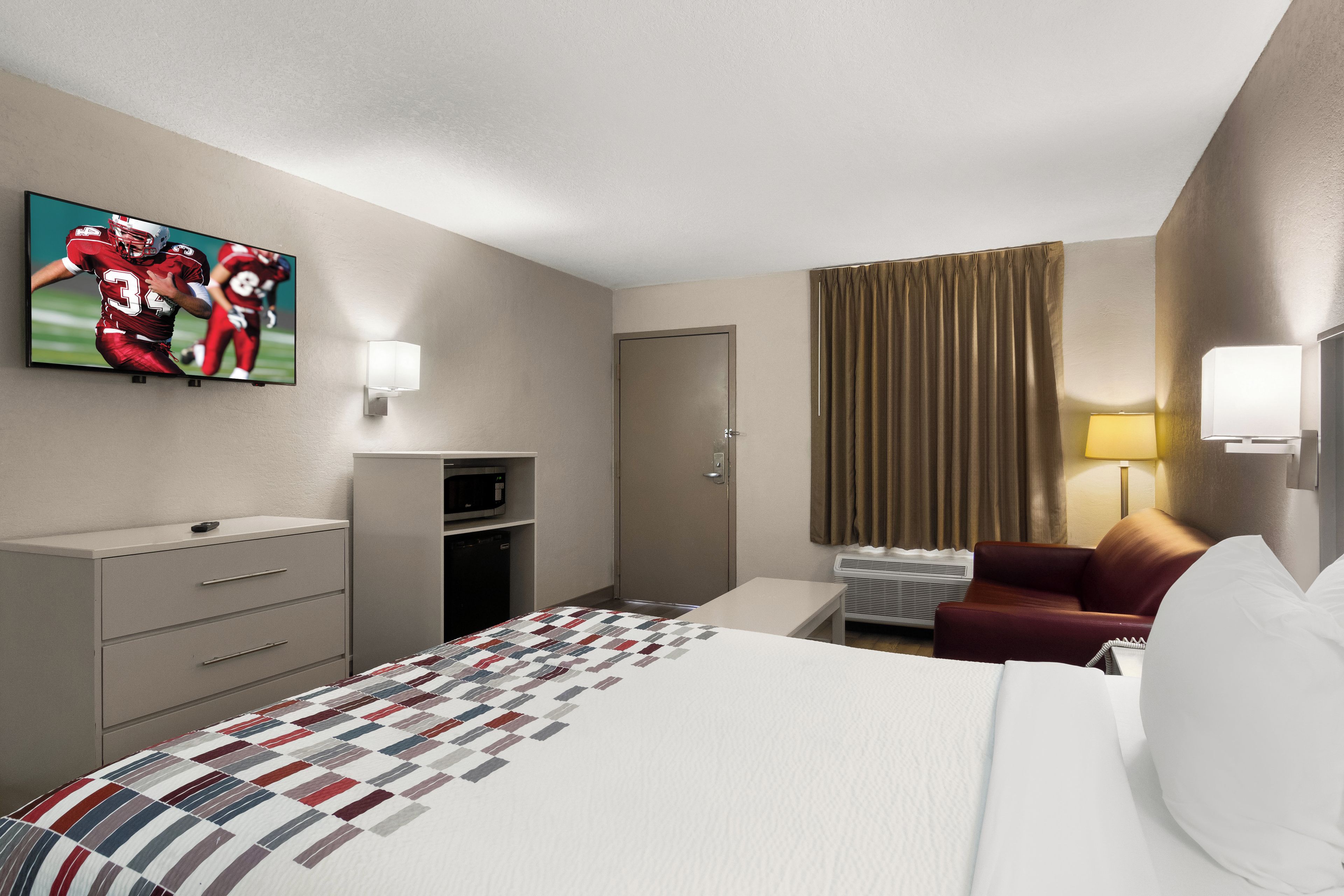 A hotel room with a bed, a television mounted on the wall, a desk with a lamp, a chair, and a door.