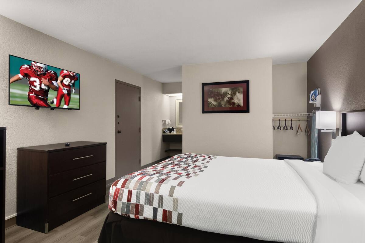 A hotel room with a large bed, a dresser, a TV on the wall, a framed picture, and a bathroom visible through an open door.