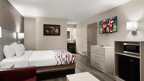 A hotel room with a bed, a desk, a chair, a TV, a mini-fridge, and a bathroom visible in the background.