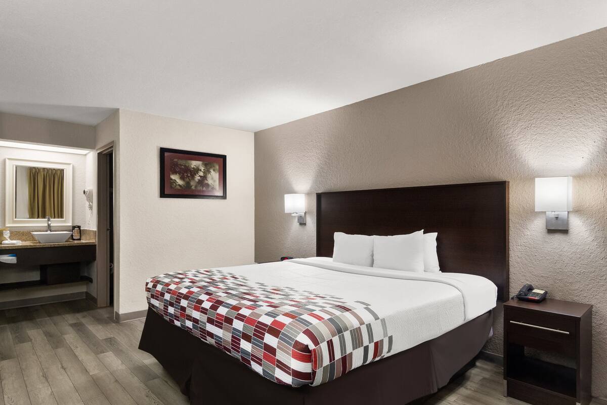A hotel room with a large bed, a bedside table with a phone, a nightstand, a wall-mounted lamp, a framed picture, and a bathroom with a sink and mirror.