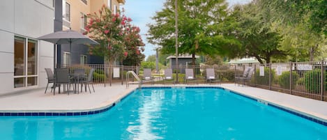 Piscina con sillas de descanso y un área de estar cubierta, rodeada de una valla y árboles.