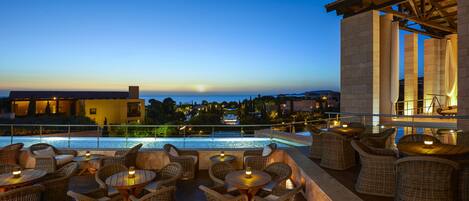 An outdoor seating area with wicker furniture and tables, overlooking a pool and a sunset view.