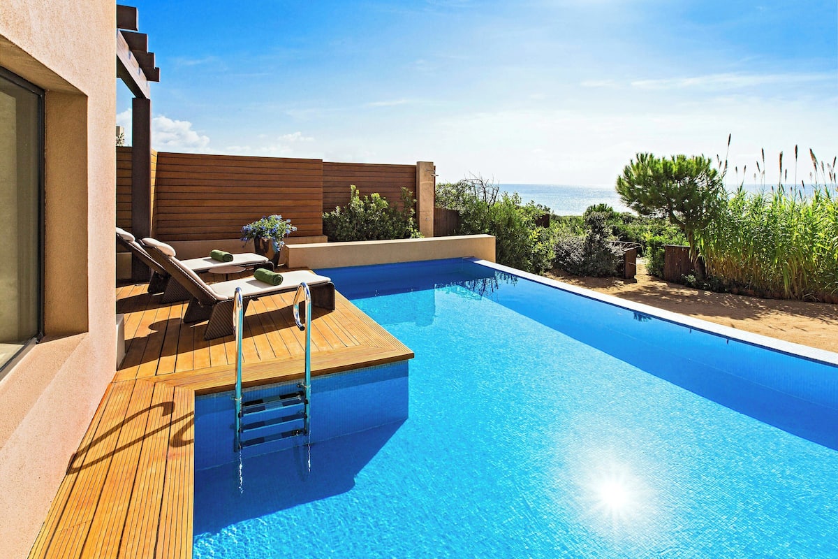 A modern outdoor swimming pool with wooden deck, lounge chairs, and a view of the sea.