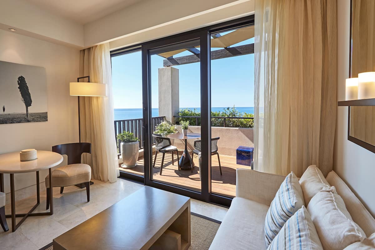 A modern hotel room with a balcony, a dining area, and a seating area with a view of the sea.