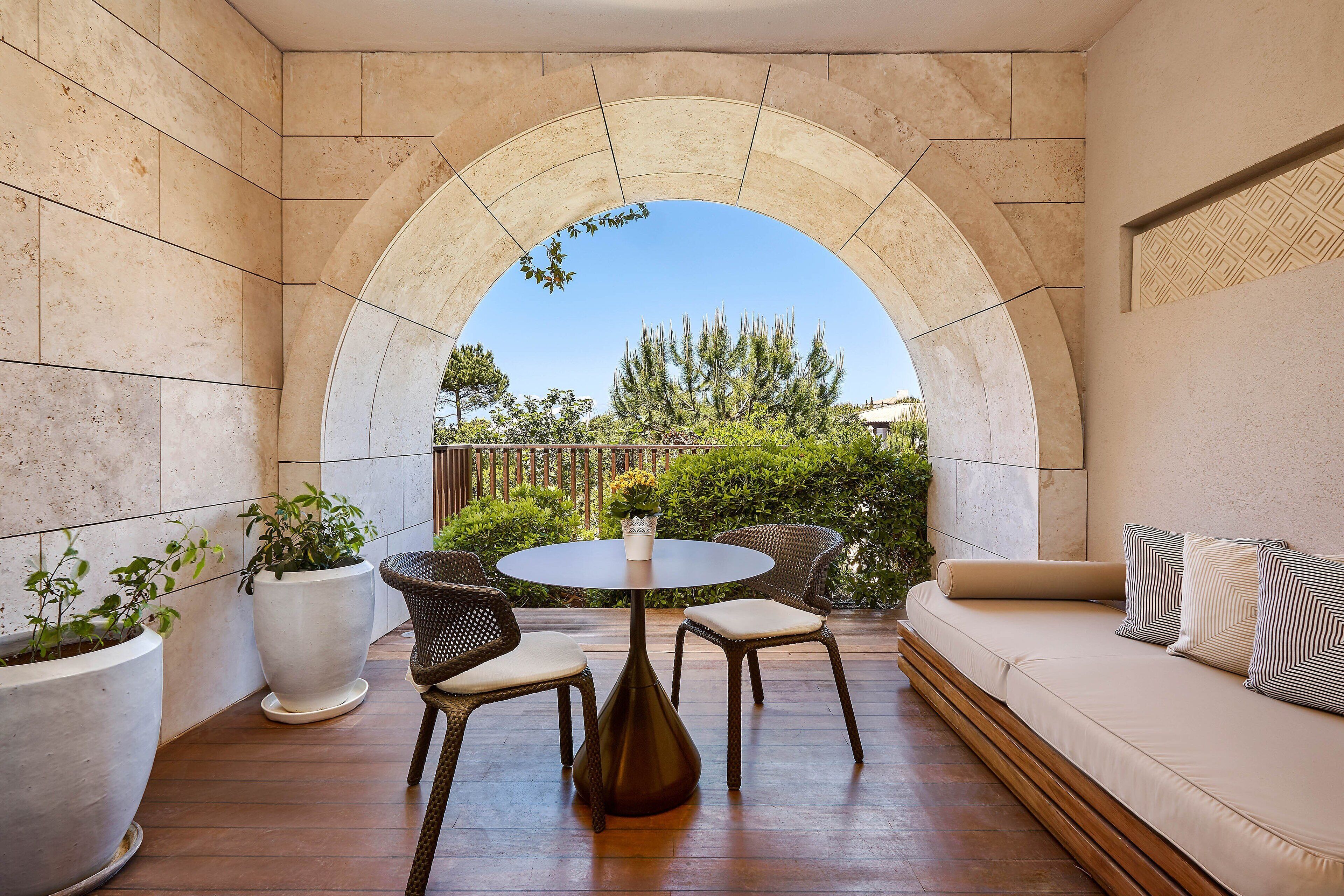 A sunlit patio with a round table and chairs, a cushioned sofa, and potted plants.