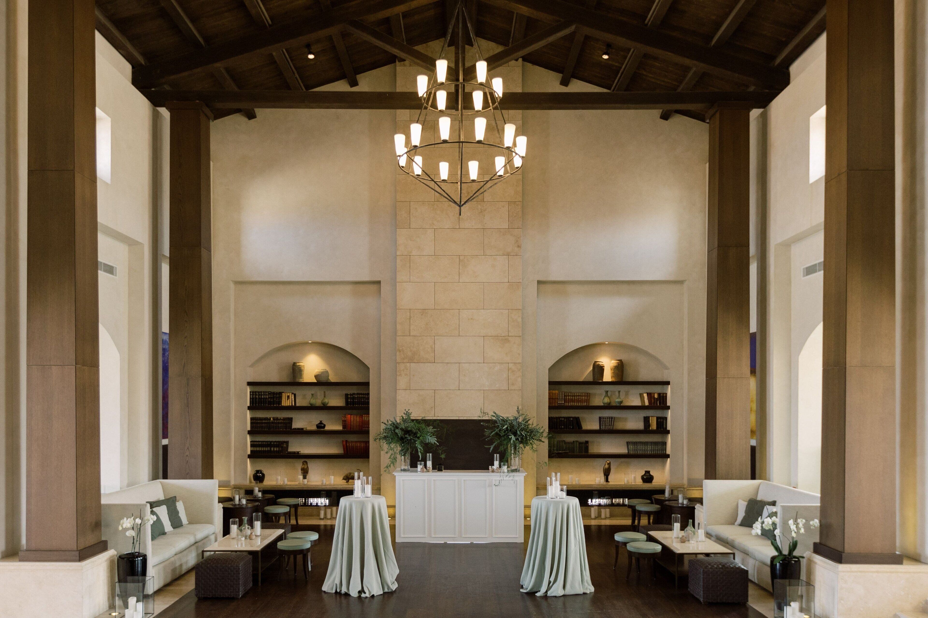A spacious lobby with a high ceiling, a central chandelier, and a fireplace. There are two sofas and a coffee table in the foreground, and bookshelves on either side.