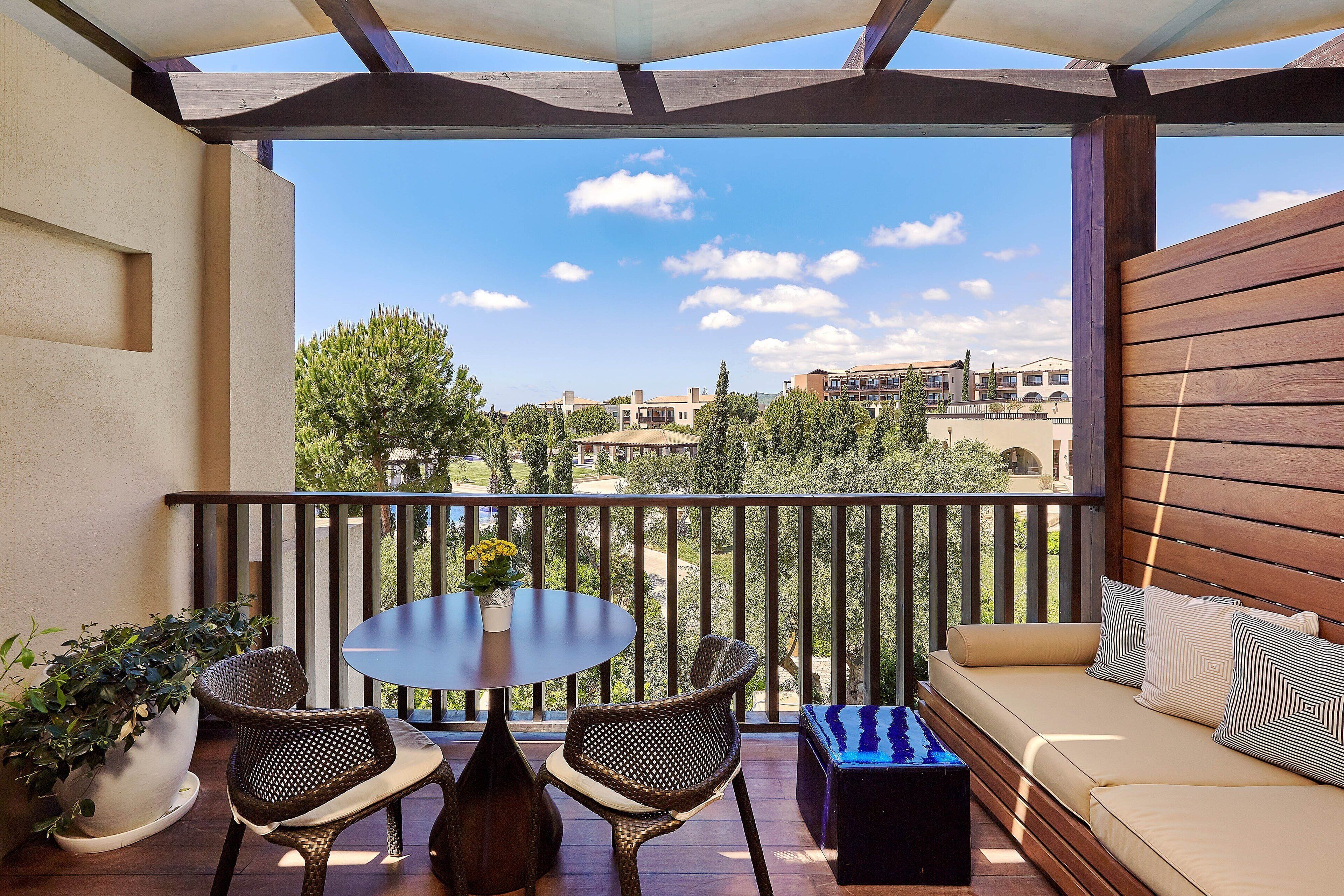 Un balcón con banco de madera, una mesa y sillas, con vista a una zona residencial con árboles y edificios.