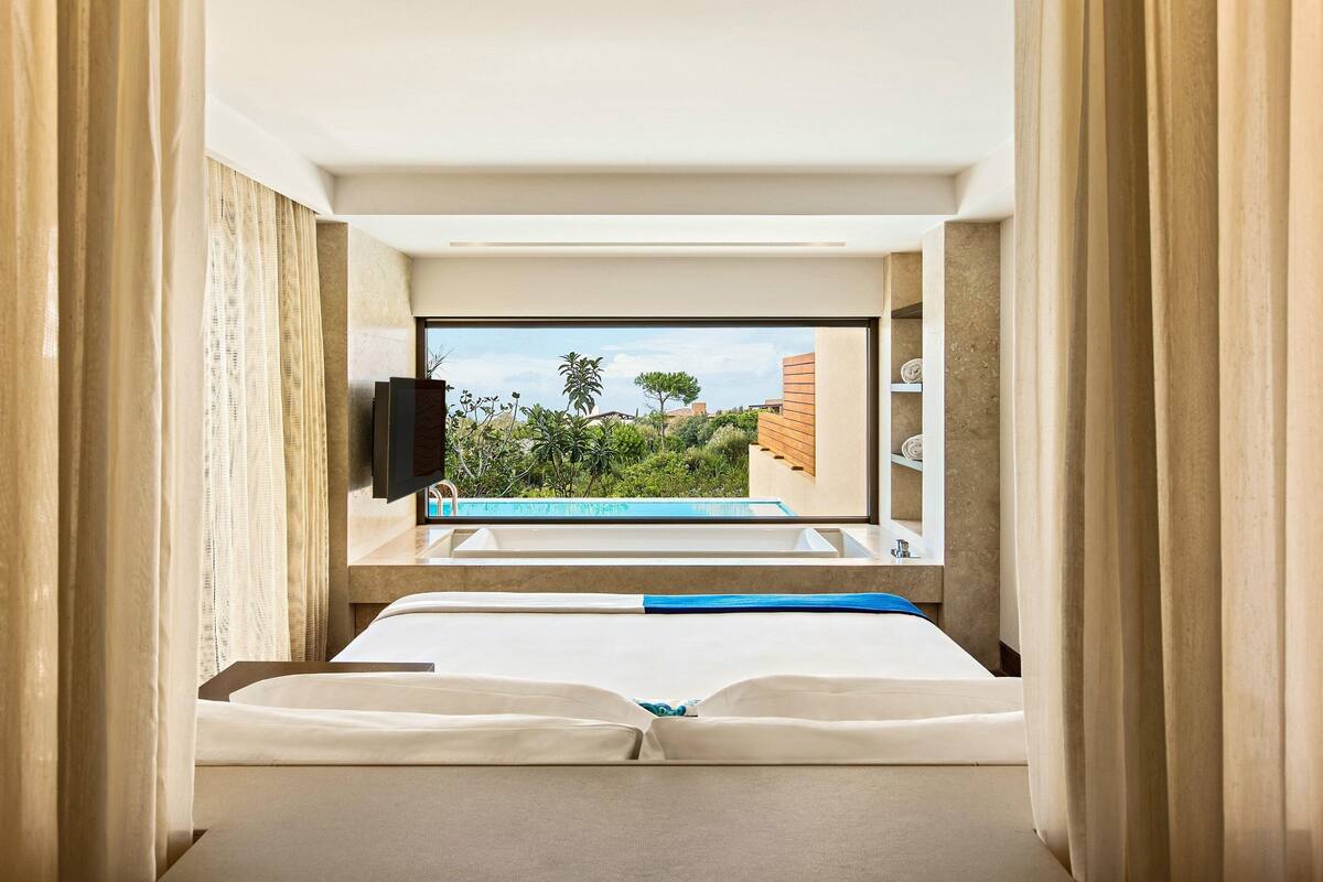 A hotel room with a bed, a TV, and a view of a swimming pool and greenery.