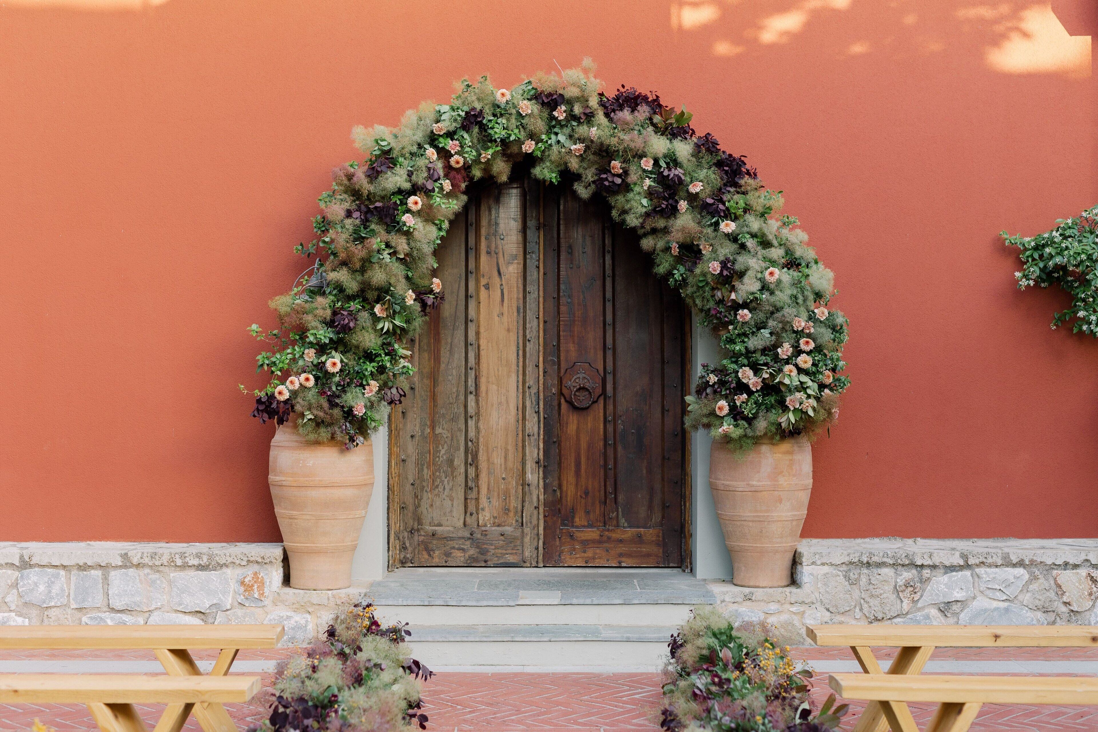 A rustic wooden door with a floral arch, flanked by large planters, set against a red wall with a stone base.