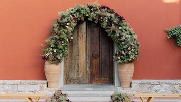 A rustic wooden door with a floral arch, flanked by large planters, set against a red wall with a stone base.