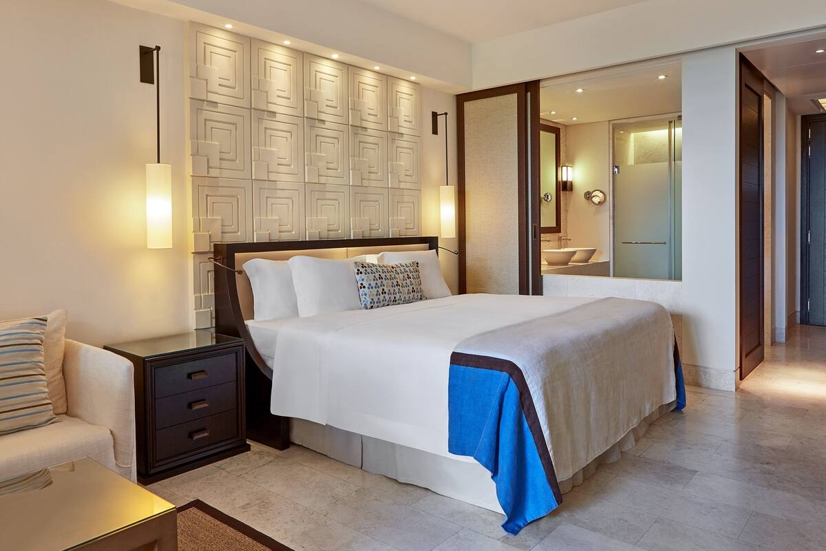 A modern hotel room with a large bed, a bedside table with drawers, a nightstand, a wall-mounted light fixture, and a bathroom visible through an open door.