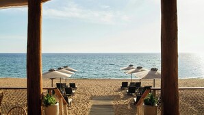 Une terrasse au bord de la plage, avec des piliers en bois, des parasols blancs et des chaises longues donnant sur l’océan.