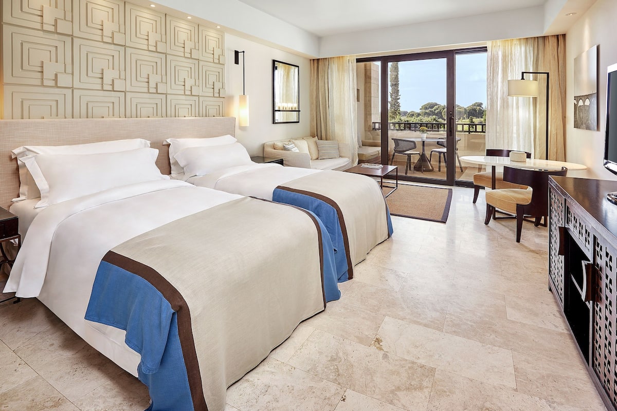 A hotel room with two beds, a desk, a chair, and a balcony with a view.