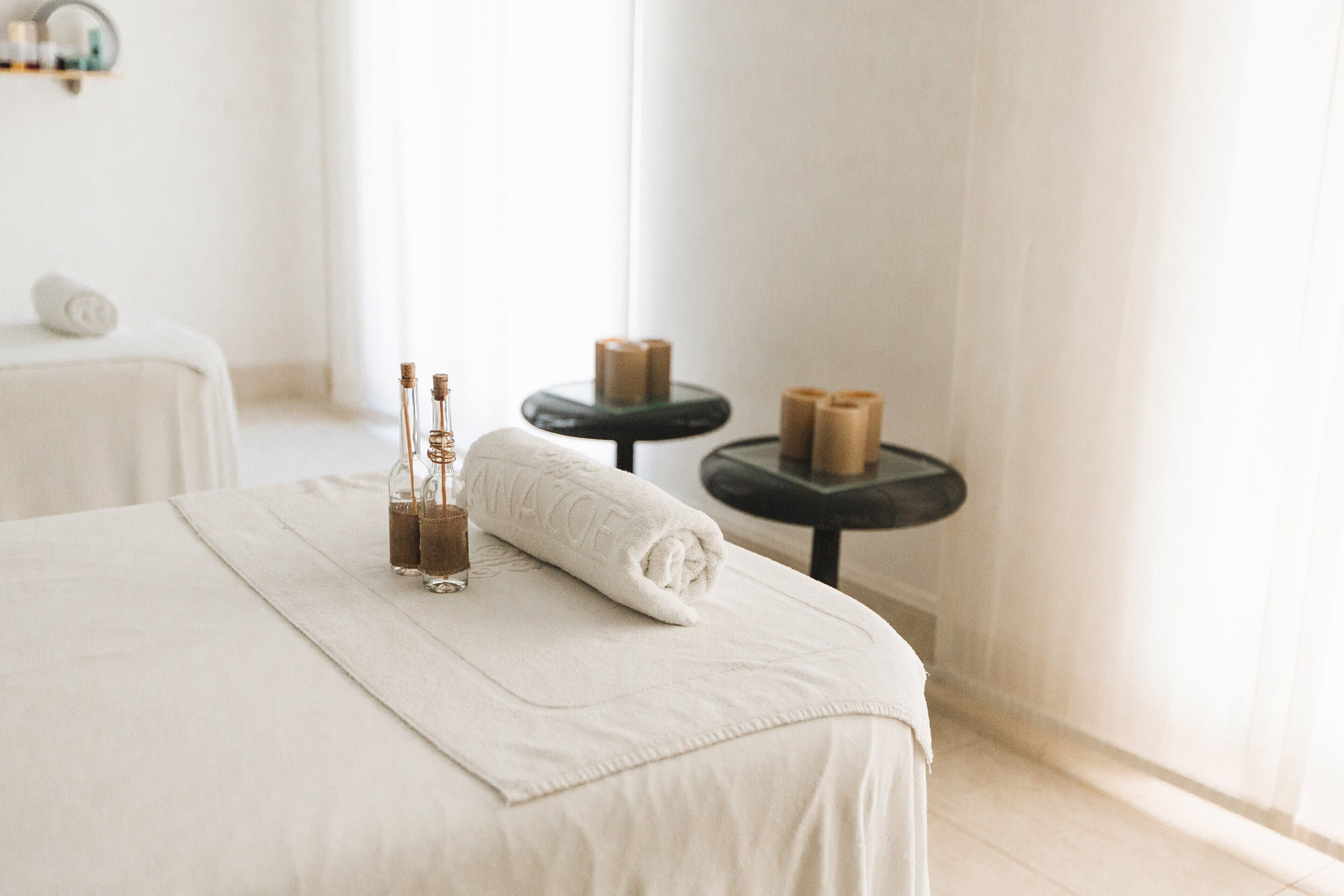 A neatly made bed with a rolled white towel and two bottles of lotion. There is a small round table with candles and a towel on it.