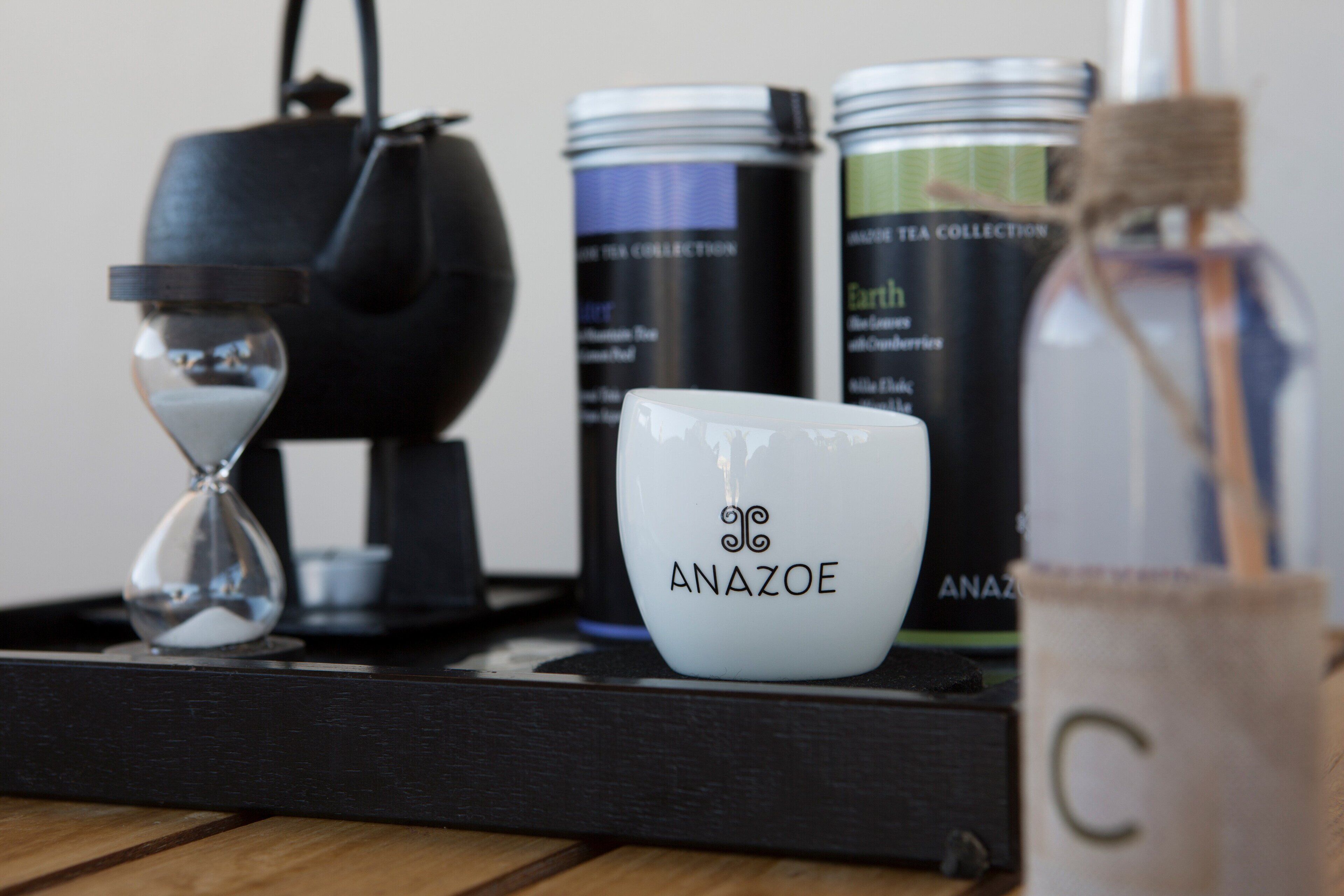Una bandeja con una taza blanca que dice "ANAZOE", una tetera negra, una ampolla de vidrio con arena y latas de té.