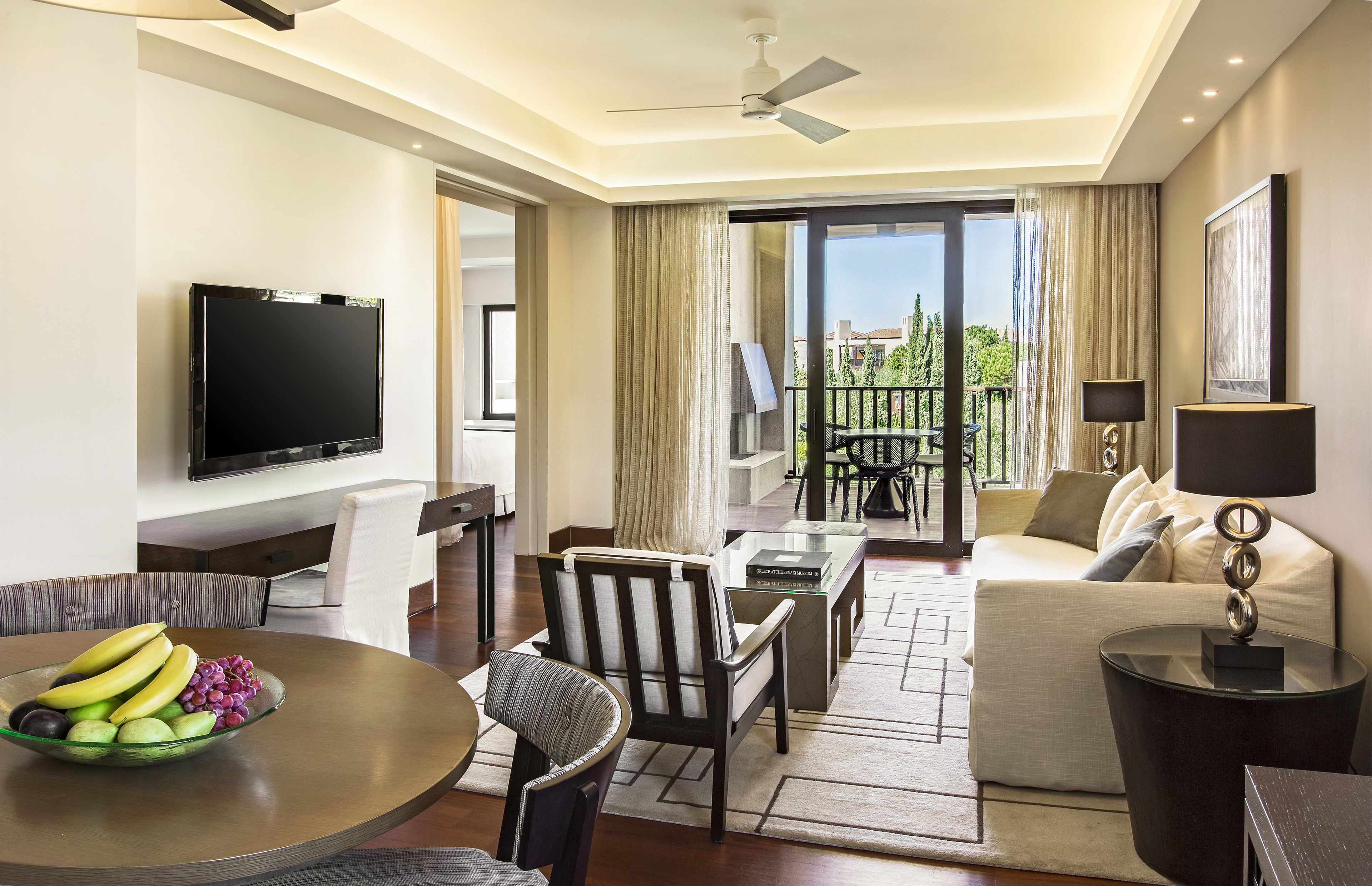 A modern hotel room with a flat-screen TV, a dining area with a fruit bowl, a sofa, and a balcony with a view.