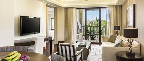 A modern hotel room with a flat-screen TV, a dining area with a fruit bowl, a sofa, and a balcony with a view.