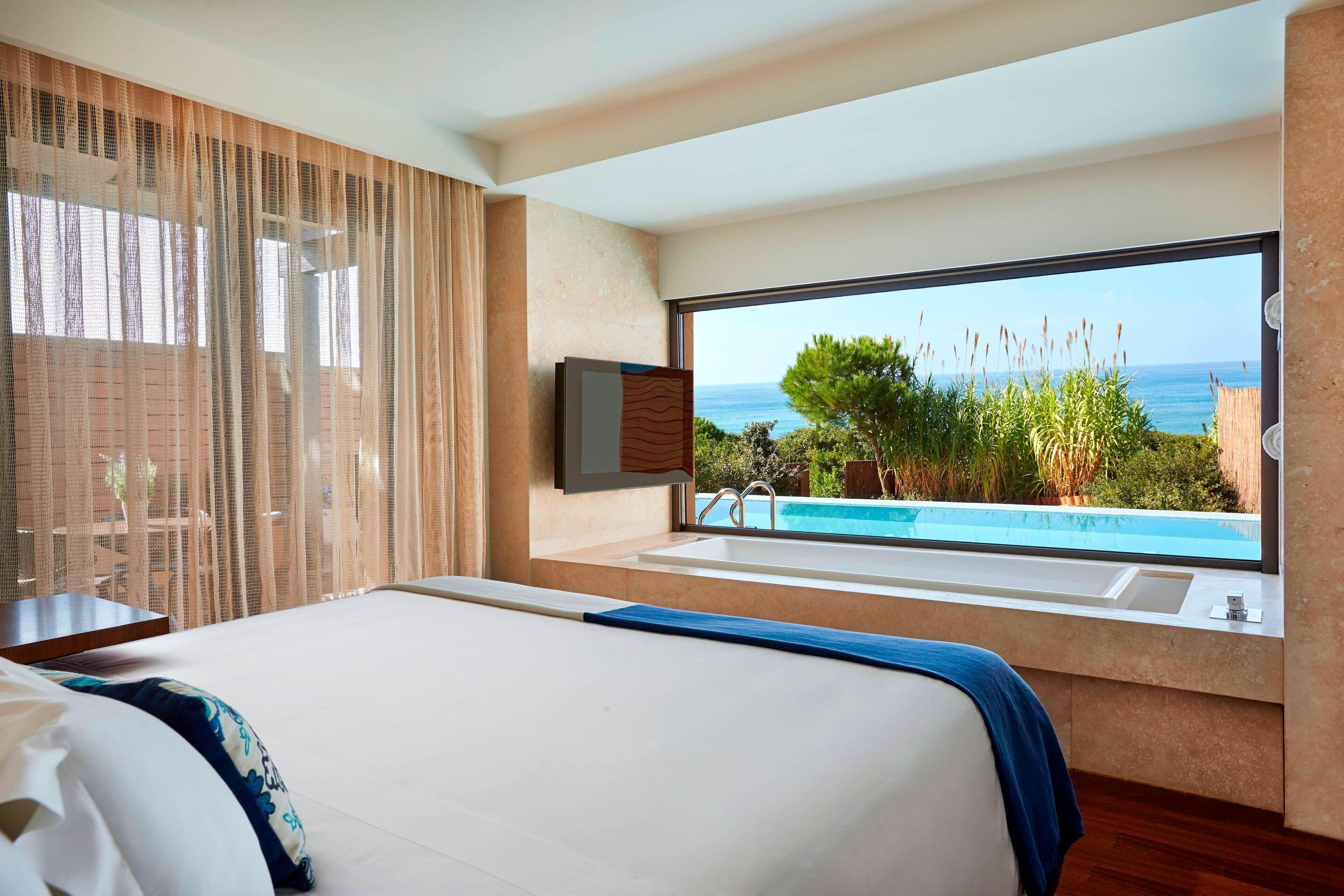 A bedroom with a large bed, a window with a view of the sea, and a bathtub.