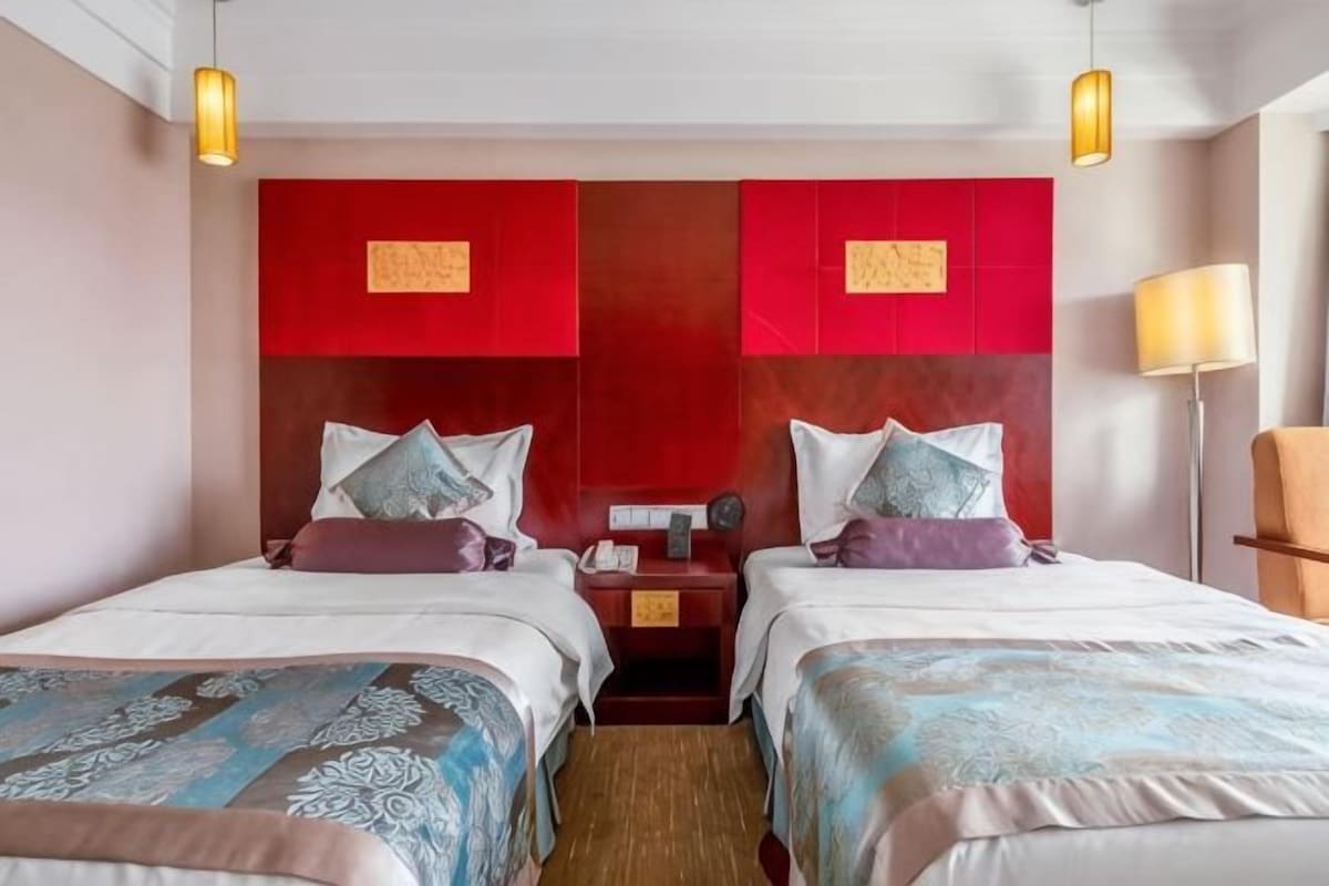 A hotel room with two beds, a red accent wall, and a nightstand with a lamp.