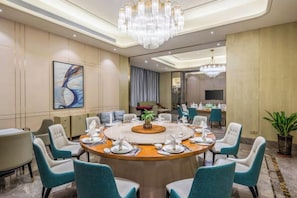 Une salle de conférence avec une table ronde préparée pour une réunion, entourée de chaises turquoise, d’un lustre et d’un grand tableau abstrait accroché au mur.