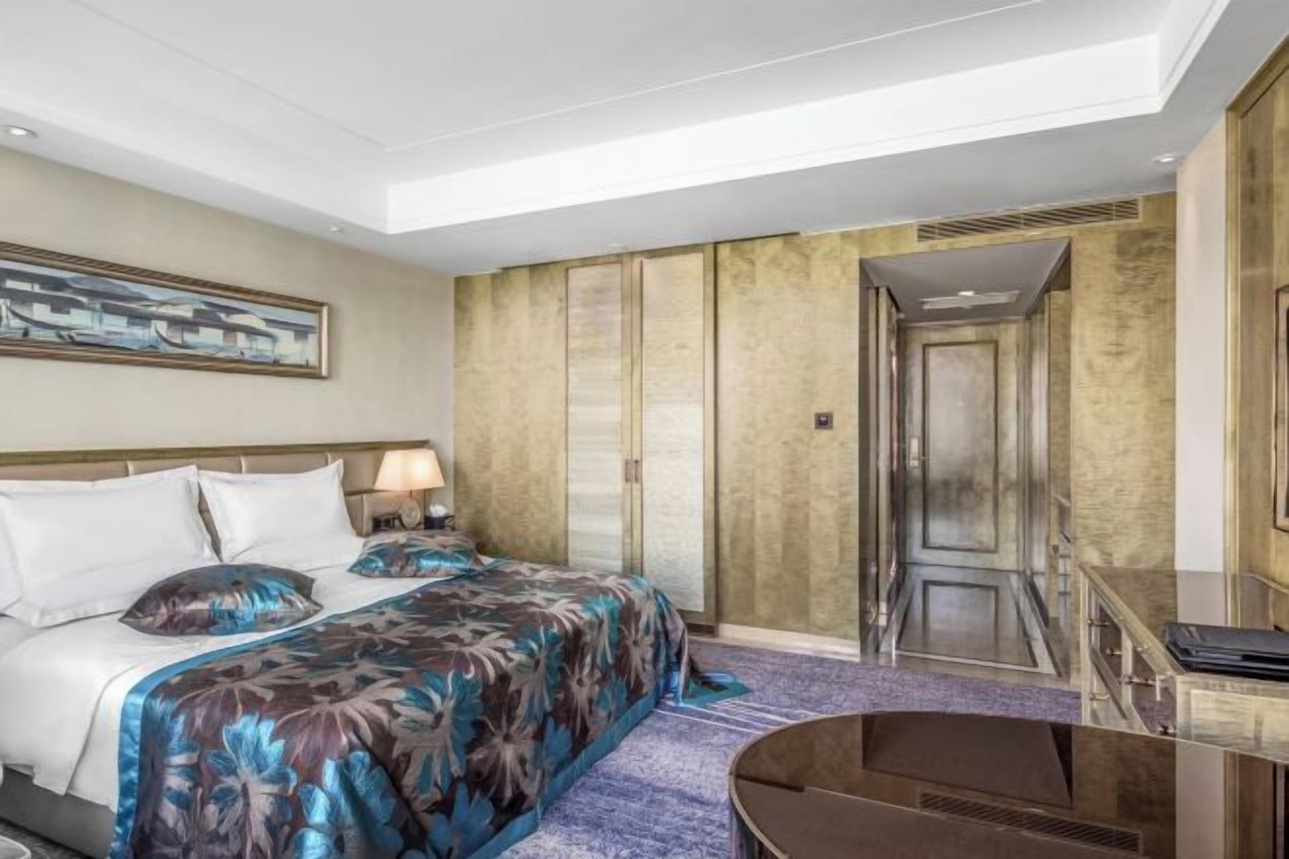 Hotellihuone, jossa on suuri sänky, työpöytä ja tuoli. Seinällä on taulu ja ovessa peili.