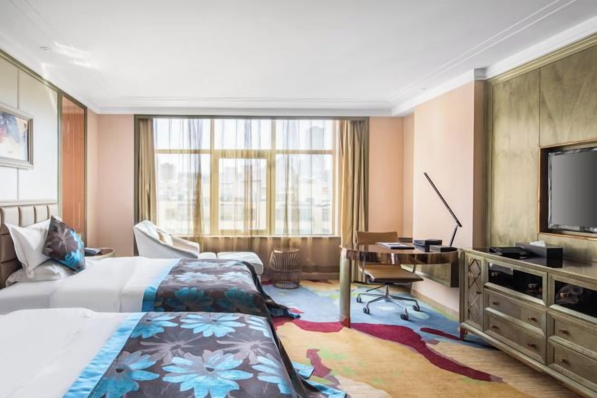 A hotel room with a bed, desk, chair, TV, and large windows.