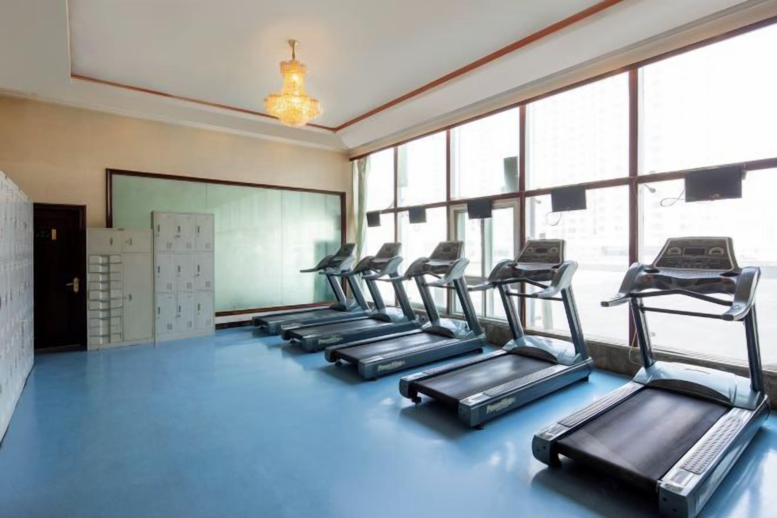 Un gimnasio con cintas de correr alineadas a lo largo de una pared, ventanas amplias y una lámpara de araña.