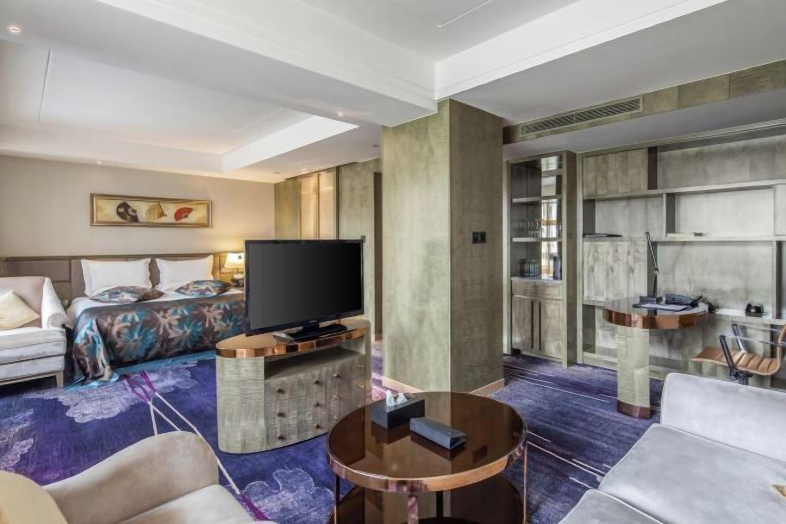 A hotel room with a bed, a television, a seating area with a coffee table, and a large window.